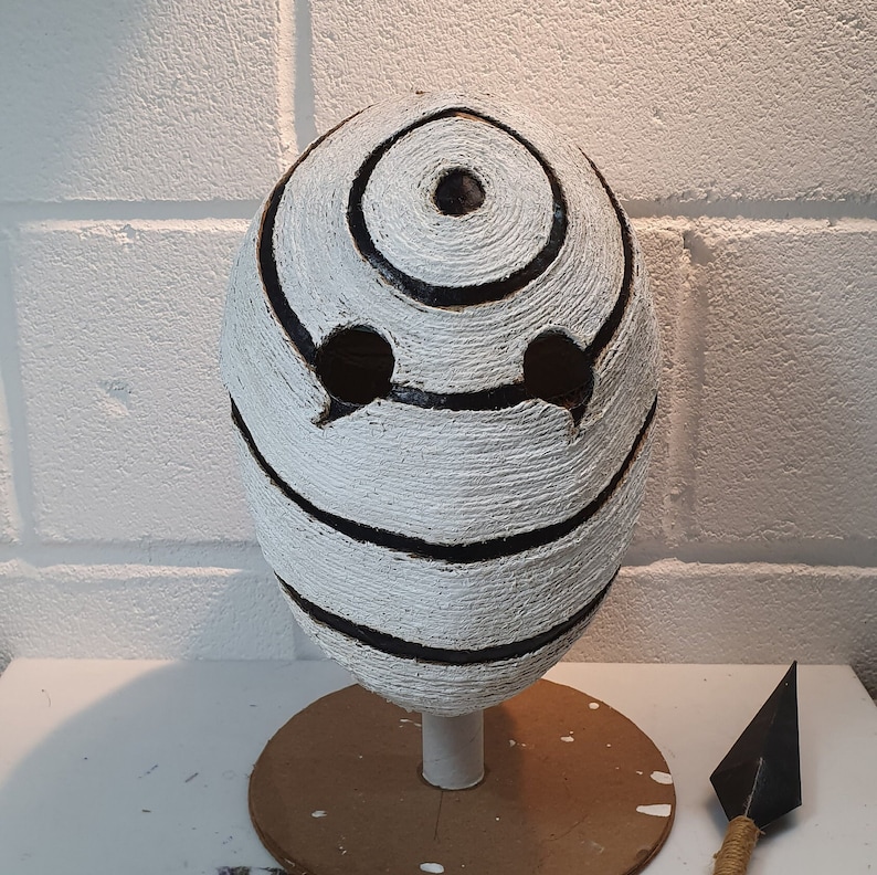 May include: White mask with black swirling patterns and two eye holes. The mask is on a brown circular stand.