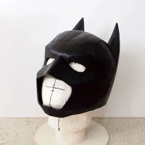 May include: A black Batman mask made of papier-m&acirc;ch&eacute;. The mask is on a white mannequin head. The mask has a pointed chin and two large ears.