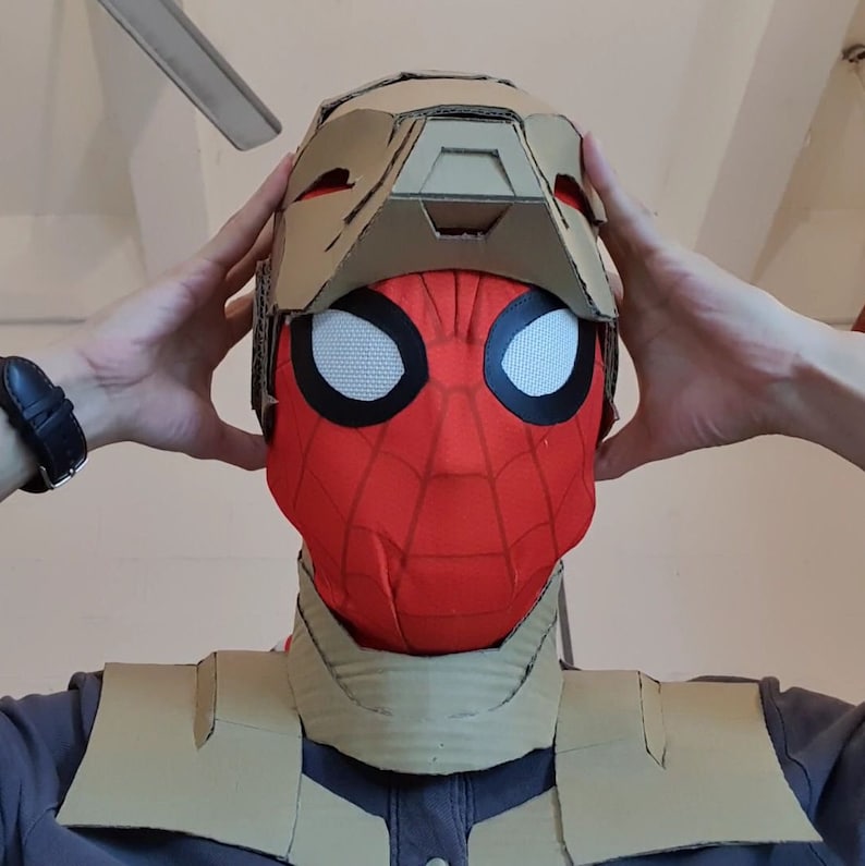 May include: A person wearing a red and black Spider-Man mask with a cardboard helmet over it. The helmet is brown and has a rectangular opening in the front. The person is also wearing a brown cardboard chest piece.