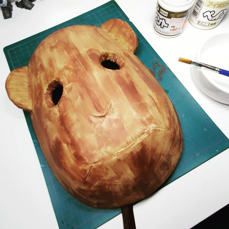 Peut inclure: Un masque en papier m&acirc;ch&eacute; brun avec deux trous pour les yeux et une petite bouche. Le masque est peint avec une peinture brun clair et brun fonc&eacute;, ce qui lui donne une texture de bois.