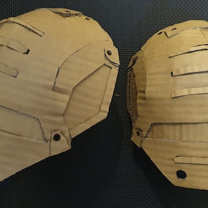 May include: Two brown cardboard pieces, possibly for a costume, with cutouts and holes. The pieces are shaped like shoulder pads.