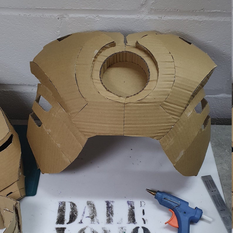 Op de afbeelding: Een kartonnen borststuk, mogelijk voor een kostuum, met een grote centrale cirkelvormige opening en hoekige zijstukken. Het stuk is gemaakt van golfkarton en toont snijlijnen en montagedetails. Een lijmpistool en liniaal zijn zichtbaar.
