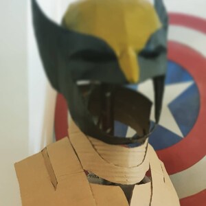 May include: A cardboard costume head and neck piece for a Wolverine cosplay. The head is yellow and black with a black mask. The neck piece is brown and has a V-shaped opening in the front.