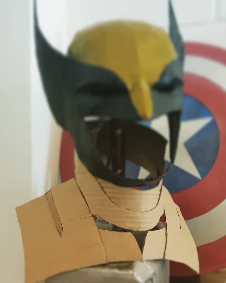May include: A cardboard costume head and neck piece for a Wolverine cosplay. The head is yellow and black with a black mask. The neck piece is brown and has a V-shaped opening in the front.