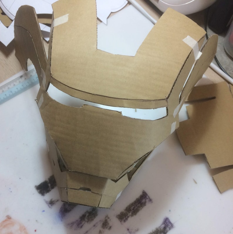 May include: A cardboard mask of Iron Man's face, with cutouts for the eyes and mouth. The mask is partially assembled and taped together.