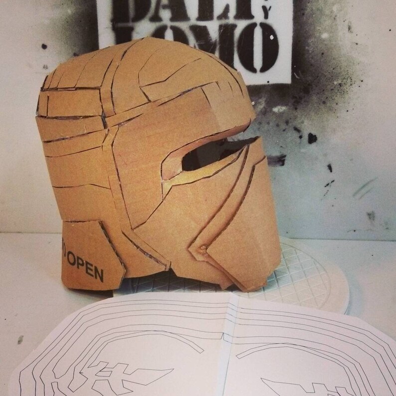 May include: A cardboard helmet with a visor, partially assembled, with the word "OPEN" printed on the side. The helmet is sitting on a white surface next to a sheet of paper with a black line drawing of a helmet.