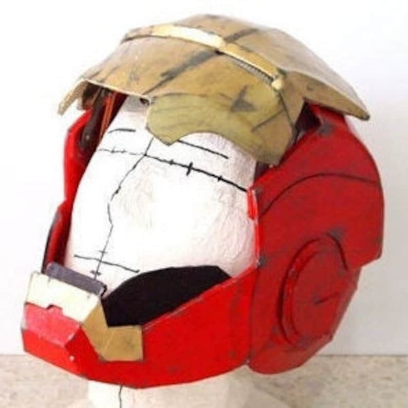 May include: A red and gold helmet with a hinged front panel. The helmet is made of cardboard and has a rough, textured surface.