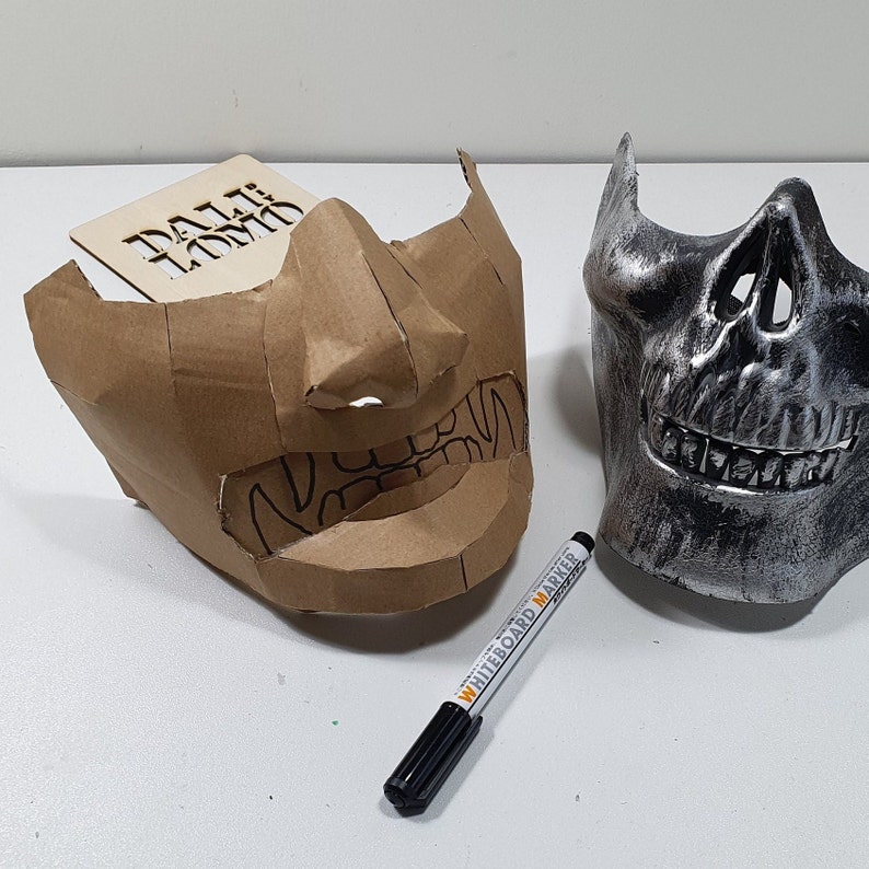 May include: Two masks, one made of brown cardboard and the other made of silver plastic. The cardboard mask has a detailed design of a face with a large nose and teeth. The silver mask has a skull design. A white marker pen is lying on the table.