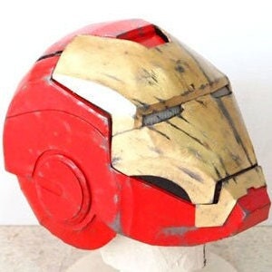 May include: A red and gold Iron Man helmet with a weathered, distressed finish. The helmet has a large, central visor and a smaller, circular visor on the side.