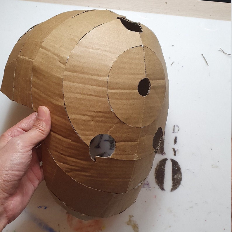 May include: A brown cardboard helmet with multiple circular cutouts. The helmet is being held by a hand.