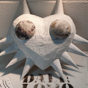 May include: A white paper mache sculpture of a heart with spikes, decorated with newspaper clippings. The sculpture is a heart with a pointed top and bottom, and the spikes are arranged around the edges. The newspaper clippings are glued to the surface of the sculpture, and they are visible through the white paint.