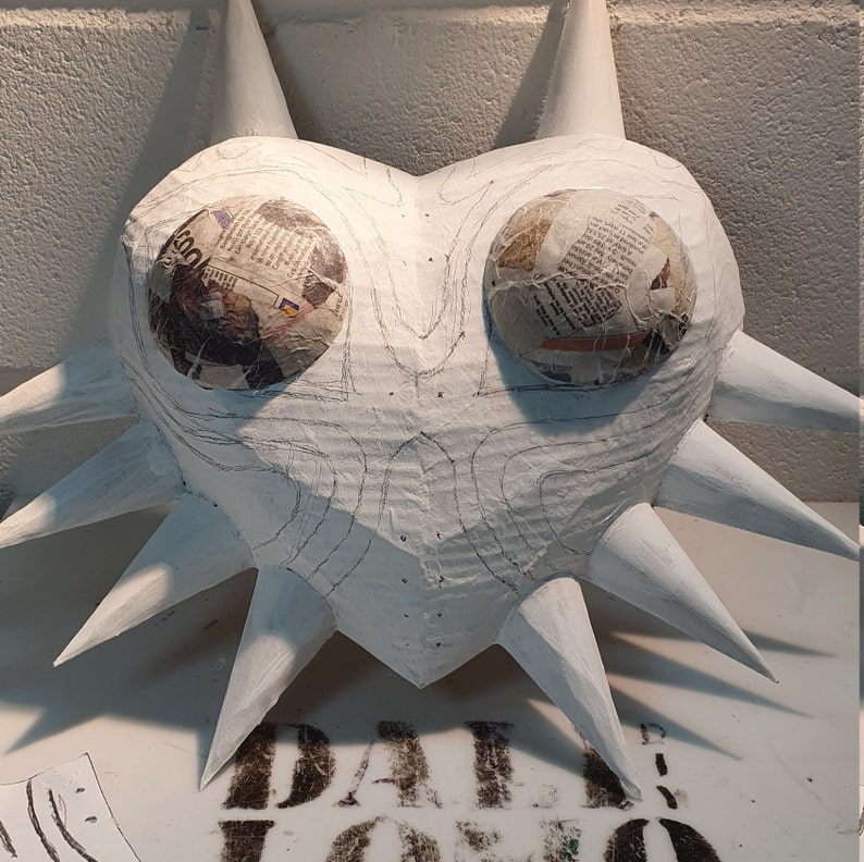 May include: A white paper mache sculpture of a heart with spikes, decorated with newspaper clippings. The sculpture is a heart with a pointed top and bottom, and the spikes are arranged around the edges. The newspaper clippings are glued to the surface of the sculpture, and they are visible through the white paint.