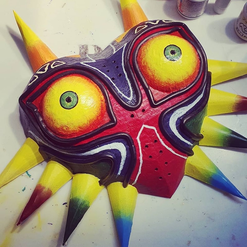 May include: A hand-painted mask with a red, yellow, and blue color scheme. The mask has a sharp, pointed design with a large, central eye and a smaller, secondary eye. The mask is made of paper mache and has a textured surface.