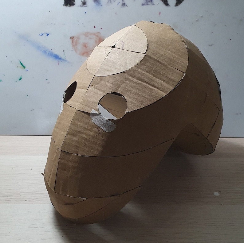 May include: A brown cardboard mask with cutouts for eyes and a mouth. The mask is shaped like a human head and has been taped together.