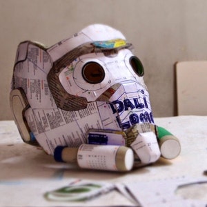 May include: A white paper mache mask with two eye holes and a green and white paper tube attached to the side. The mask is partially covered in printed paper with the word "DALI" visible.