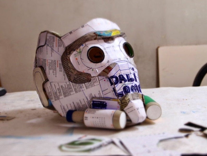 May include: A white paper mache mask with two eye holes and a green and white paper tube attached to the side. The mask is partially covered in printed paper with the word "DALI" visible.