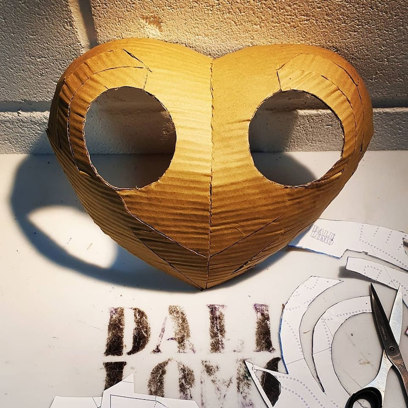 May include: A brown cardboard mask shaped like a heart with two eye holes cut out. The mask is sitting on a white surface with paper patterns and a pair of scissors.