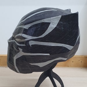 May include: Black and silver paper mache mask of a superhero with a detailed design. The mask is on a black stand.