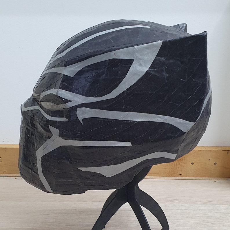 May include: Black and silver paper mache mask of a superhero with a detailed design. The mask is on a black stand.