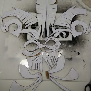 May include: A white paper stencil of a fleur-de-lis design with multiple pieces. The stencil is marked with letters L and R for left and right.