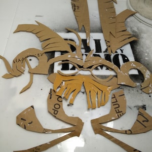 May include: A set of brown cardboard stencils for creating a mask or design. The stencils are cut out in the shape of a face with eyes, nose, and mouth.