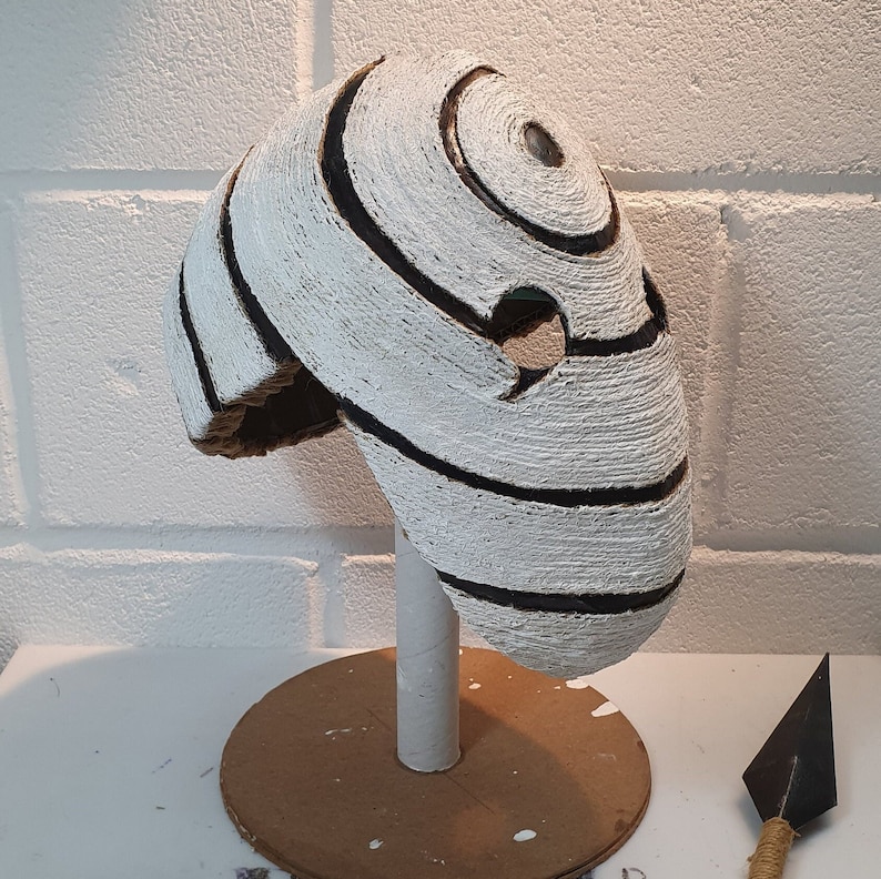 May include: White and black mask with a spiral design. The mask is made of cardboard and is on a cardboard stand.
