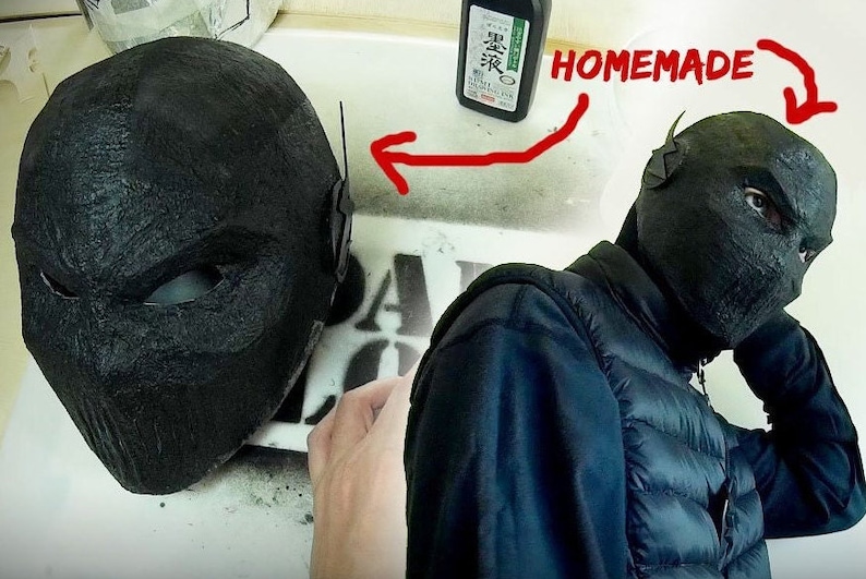 May include: A black and gray mask with a textured surface. The mask has a lightning bolt design on the side. The mask is being held by a person wearing a black jacket. The text "HOMEMADE" is written in red on a white surface.