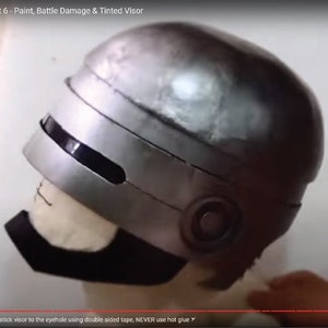 Può includere: Un casco argentato con una visiera nera e un cinturino per il mento nero. Il casco è fatto di un materiale duro e ha una superficie liscia. La visiera è colorata e ha una cornice nera. Il cinturino per il mento è fatto di un materiale morbido e ha una fibbia.