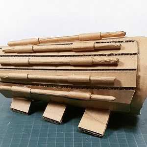 May include: A brown cardboard arm guard with multiple layers of cardboard tubes attached to the front. The arm guard has a rounded shape and is designed to protect the wearer's arm.