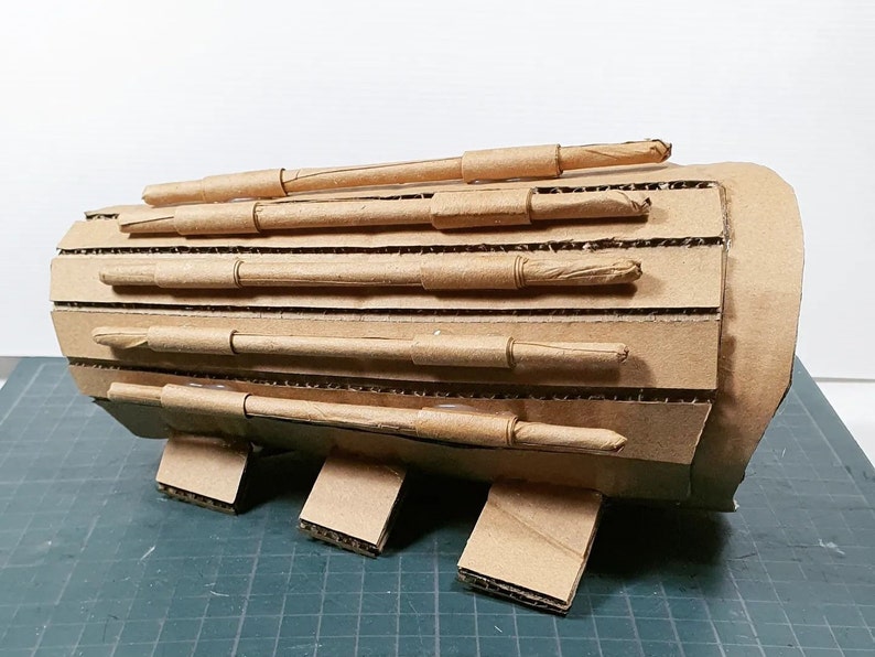 May include: A brown cardboard arm guard with multiple layers of cardboard tubes attached to the front. The arm guard has a rounded shape and is designed to protect the wearer's arm.