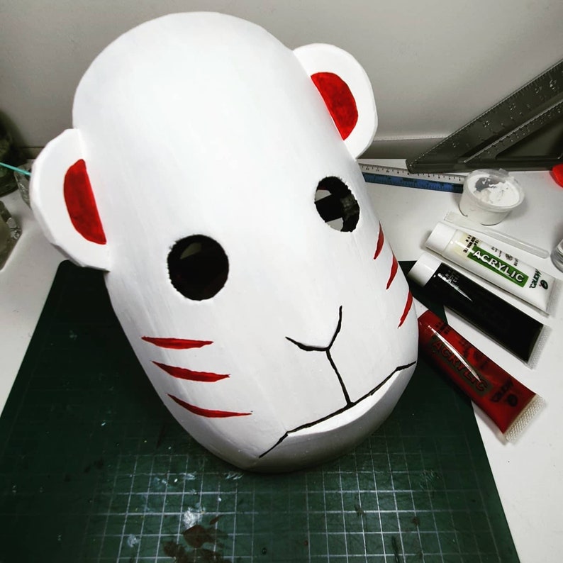 Peut inclure: Masque blanc avec des d&eacute;tails rouges, y compris les oreilles et les rayures. Le masque est sur un tapis de coupe vert avec des tubes de peinture rouge, noir et blanc.