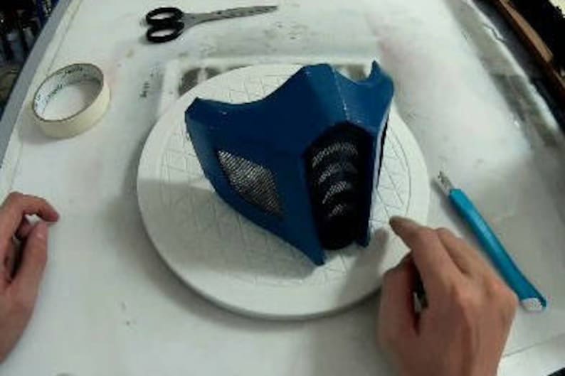May include: A blue plastic mask with a black mesh covering the eyes. The mask is sitting on a white circular surface.
