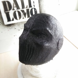 May include: A black, textured, and detailed mask made of papier-m&acirc;ch&eacute;. The mask is shaped like a human head and has a closed mouth and eyes.