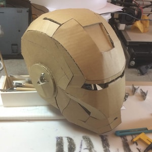 May include: A cardboard Iron Man helmet, partially assembled, with visible details like the eye slits and the chin piece. The helmet is resting on a white surface.