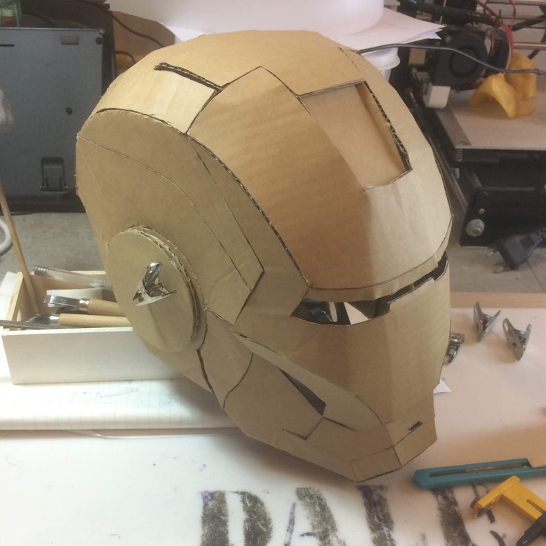 May include: A cardboard Iron Man helmet, partially assembled, with visible details like the eye slits and the chin piece. The helmet is resting on a white surface.