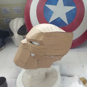 May include: A brown cardboard mask with a detailed design, resembling a superhero's mask. The mask is mounted on a white plaster head form.