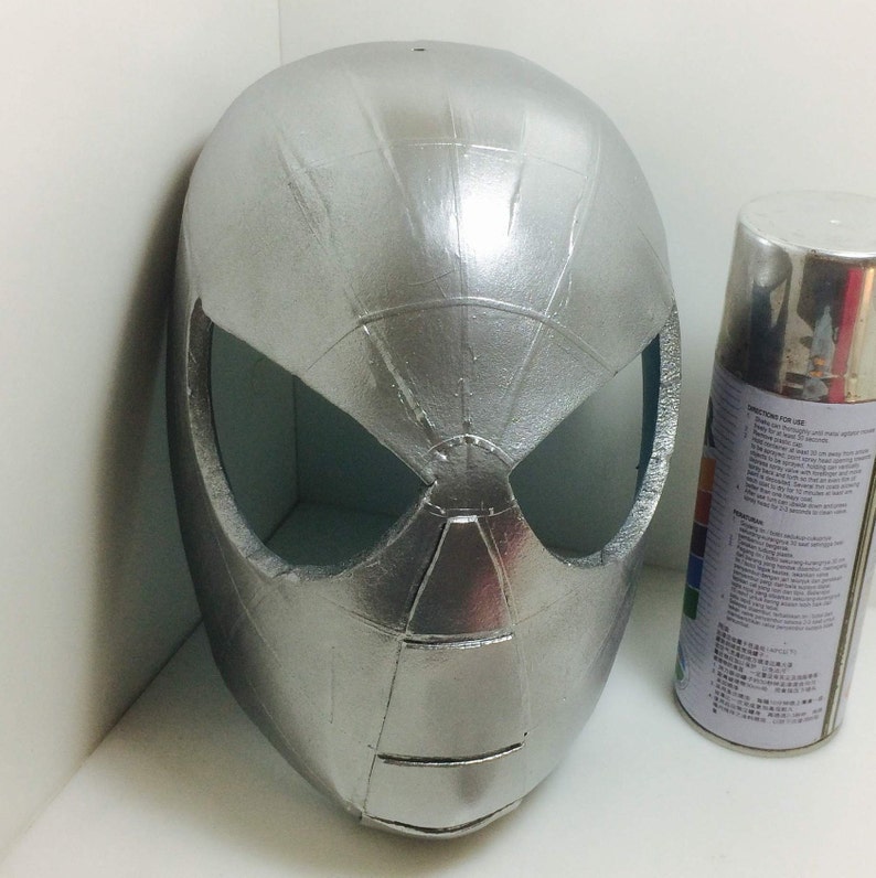 May include: A silver-colored plastic mask with a spider-web design. The mask has two eye holes and a small hole on top. The mask is sitting on a white surface next to a can of silver spray paint.