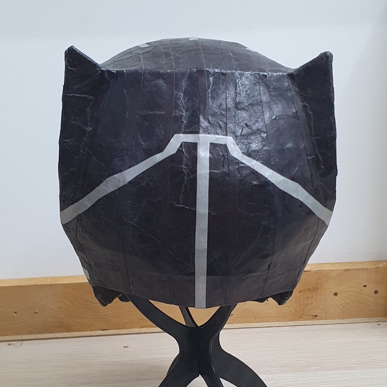 May include: Black paper mache mask with a white design resembling a panther's face. The mask is on a black stand.