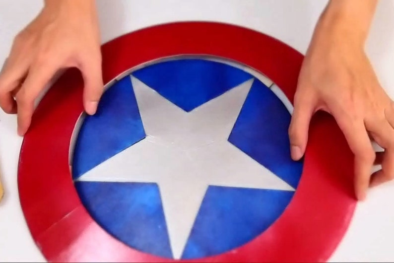 May include: A red, white, and blue cardboard shield with a five-pointed star in the center. The shield is being held by two hands.