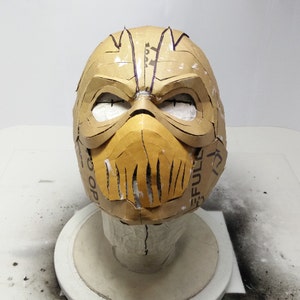 May include: A brown cardboard mask with a detailed design. The mask has a raised, textured, and segmented lower face. The mask is on a white surface.