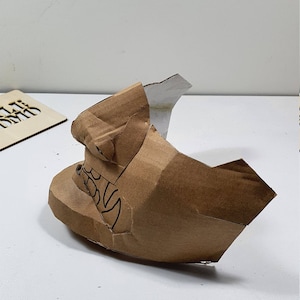 May include: A brown cardboard mask with a white paper nose and a black design on the side. The mask is partially folded and appears to be in the process of being made.