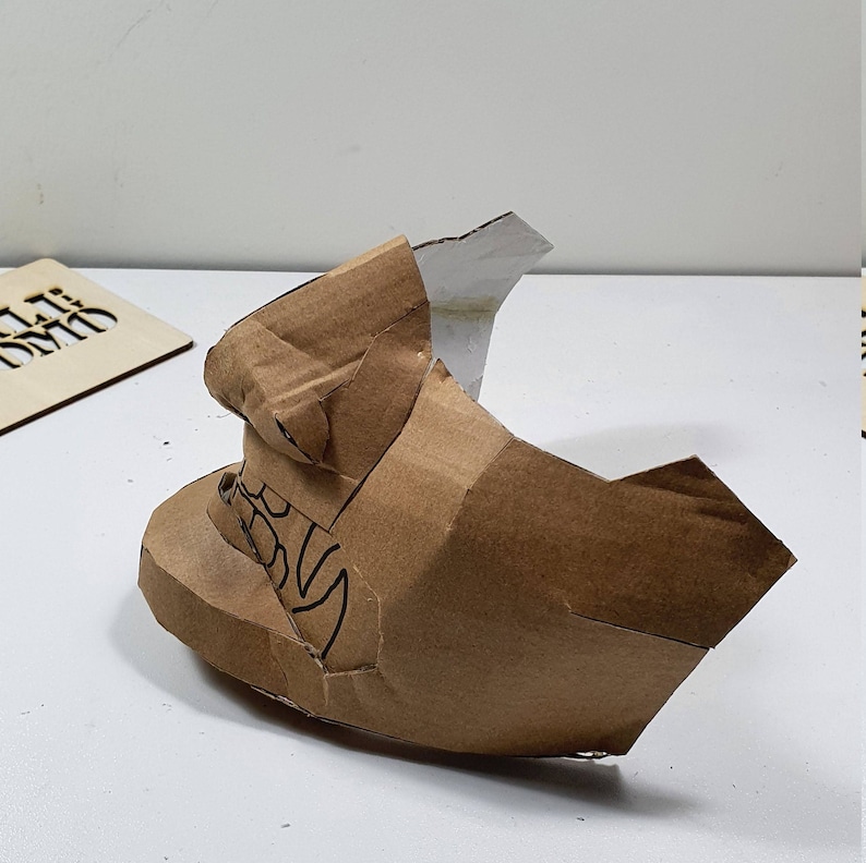 May include: A brown cardboard mask with a white paper nose and a black design on the side. The mask is partially folded and appears to be in the process of being made.