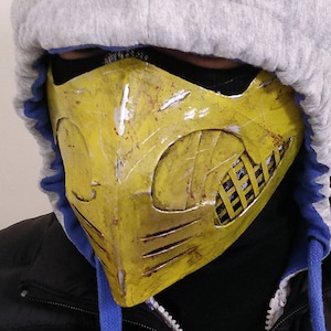 May include: A yellow and black face mask with a textured surface and a grid pattern on the right side. The mask is worn by a person wearing a grey hooded sweatshirt.