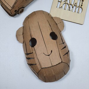 Peut inclure: Un masque en carton brun avec deux yeux noirs, un petit sourire et des marques ray&eacute;es sur les c&ocirc;t&eacute;s. Le masque est fait de carton et a une surface textur&eacute;e.
