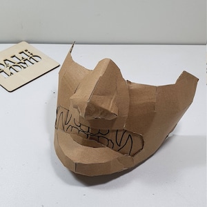 May include: A brown cardboard mask with a detailed design of a face with teeth and a nose. The mask is made of multiple pieces of cardboard that have been glued together.