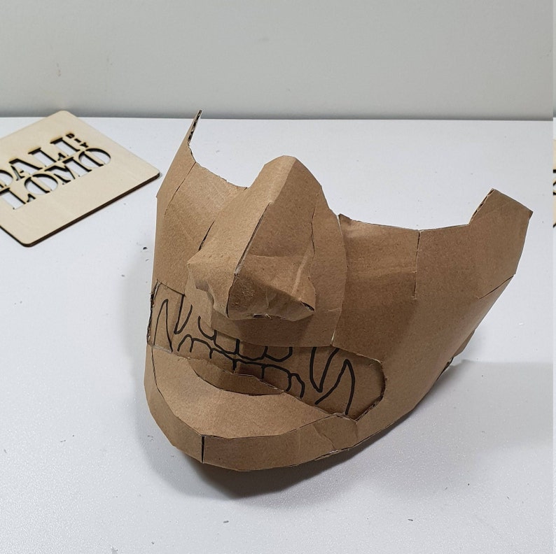 May include: A brown cardboard mask with a detailed design of a face with teeth and a nose. The mask is made of multiple pieces of cardboard that have been glued together.