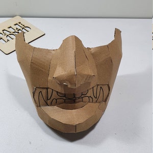 May include: A brown cardboard mask with a detailed design of a face with sharp teeth and a prominent nose.