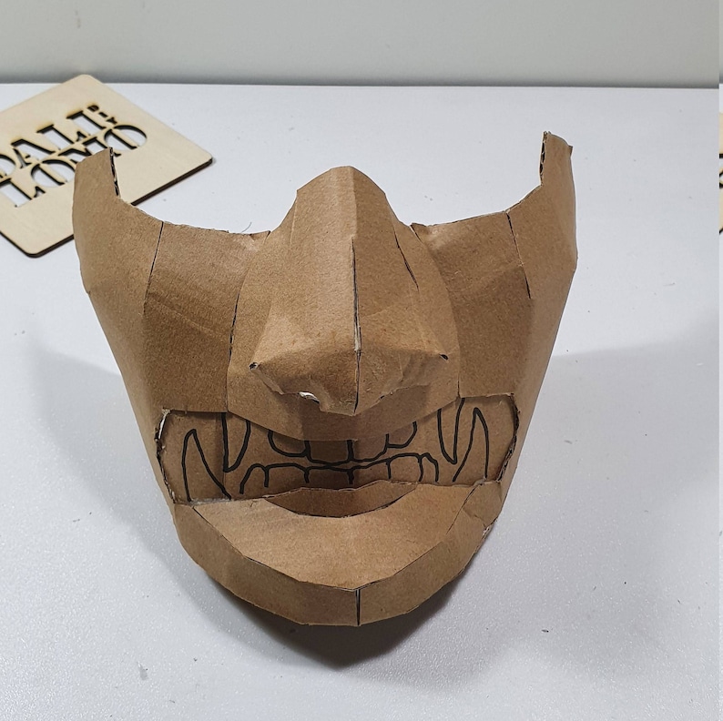 May include: A brown cardboard mask with a detailed design of a face with sharp teeth and a prominent nose.
