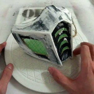 May include: A white and black mask with a green mesh covering the eyes. The mask has a textured surface and is partially painted black.