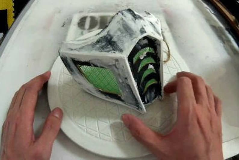 May include: A white and black mask with a green mesh covering the eyes. The mask has a textured surface and is partially painted black.